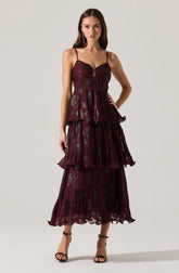A woman is wearing a dark red, tiered, pleated dress with metallic floral accents. The dress has thin spaghetti straps and a fitted bodice. She pairs it with black high-heeled sandals that have ankle straps.